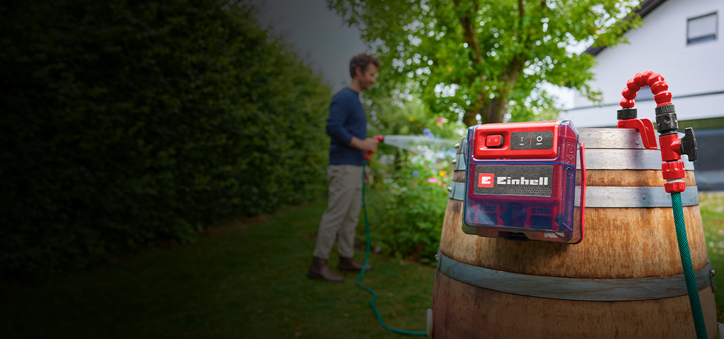 Einhell Akku-Klarwasserpumpe an einem Holzfass im Garten, während eine Person im Hintergrund Pflanzen bewässert.