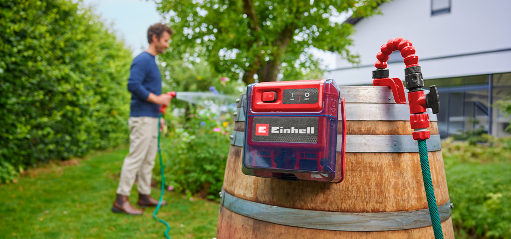 Einhell Akku-Klarwasserpumpe an einem Holzfass im Garten, während eine Person im Hintergrund Pflanzen bewässert.
