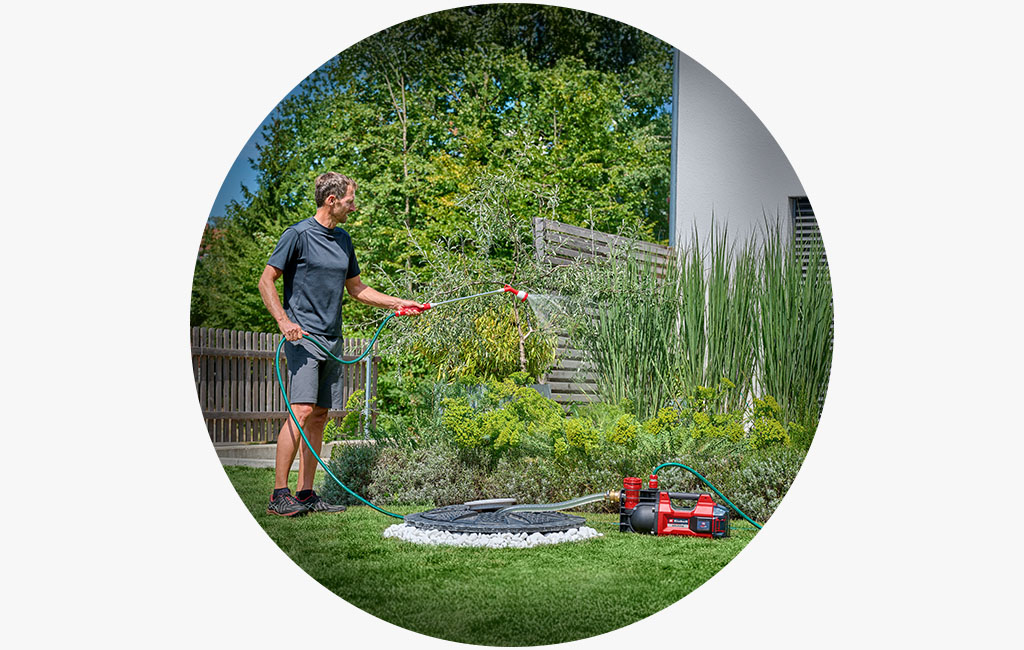 Ein Mann bewässert mit einer Einhell Akku-Gartenpumpe einen Vorgarten mit vielen grünen Pflanzen vor einem Haus.