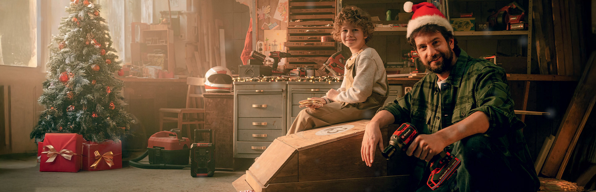 Ein Mann mit Weihnachtsmütze und Akkuschrauber sitzt mit einem Kind in einer Werkstatt neben einem selbstgebauten Holzauto. Im Hintergrund stehen ein geschmückter Weihnachtsbaum und rote Geschenke.