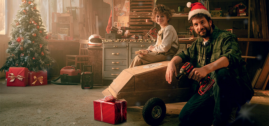 Ein Mann mit Weihnachtsmütze und Akkuschrauber sitzt mit einem Kind in einer Werkstatt neben einem selbstgebauten Holzauto. Im Hintergrund stehen ein geschmückter Weihnachtsbaum und rote Geschenke.
