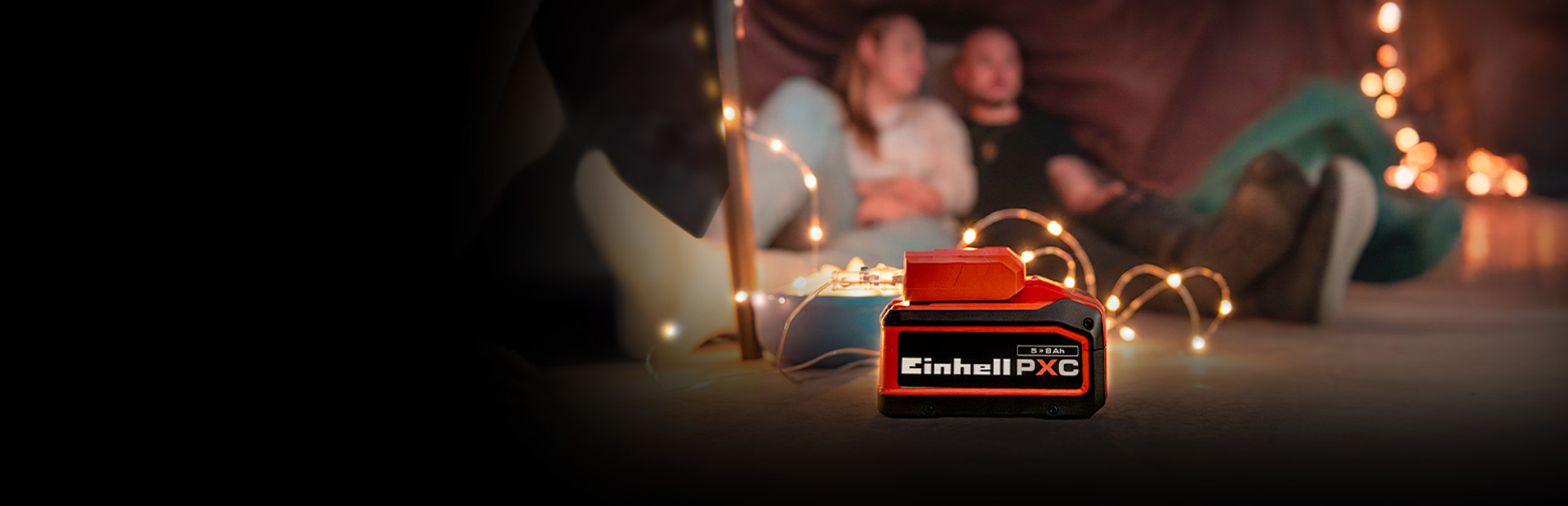Ein Einhell Power X-Change Akku (5,2 Ah) steht im Vordergrund, im Hintergrund sitzt ein Paar in gemütlicher Atmosphäre mit Lichterkette.