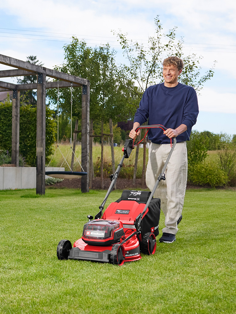 A man mows a lawn with the red Einhell cordless lawn mower GC-CM 36/51 S Li BL in front of a garden area with a wooden pergola.
