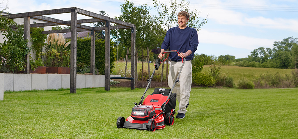 A man mows a lawn with the red Einhell cordless lawn mower GC-CM 36/51 S Li BL in front of a garden area with a wooden pergola.