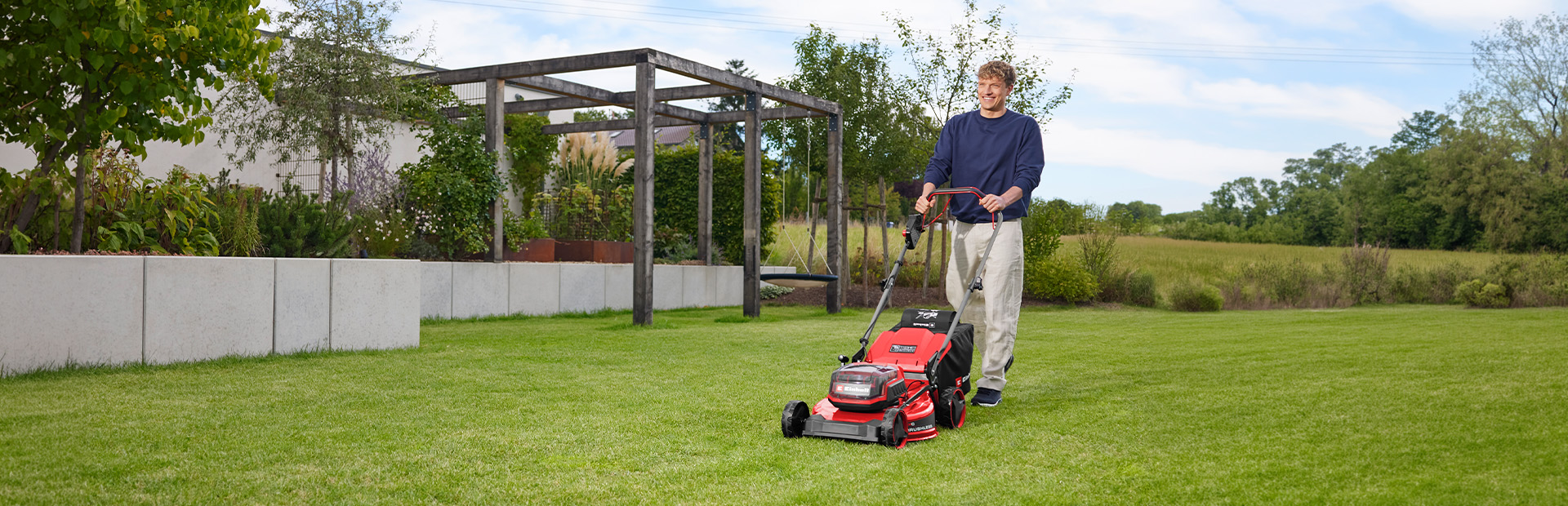 A man mows a lawn with the red Einhell cordless lawn mower GC-CM 36/51 S Li BL in front of a garden area with a wooden pergola.