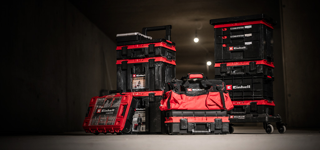 Mehrere rote Einhell E-Case Werkzeugkoffer mit schwarzen Details, teils gestapelt, sowie eine rote Werkzeugtasche in einer dunklen Werkstatt.