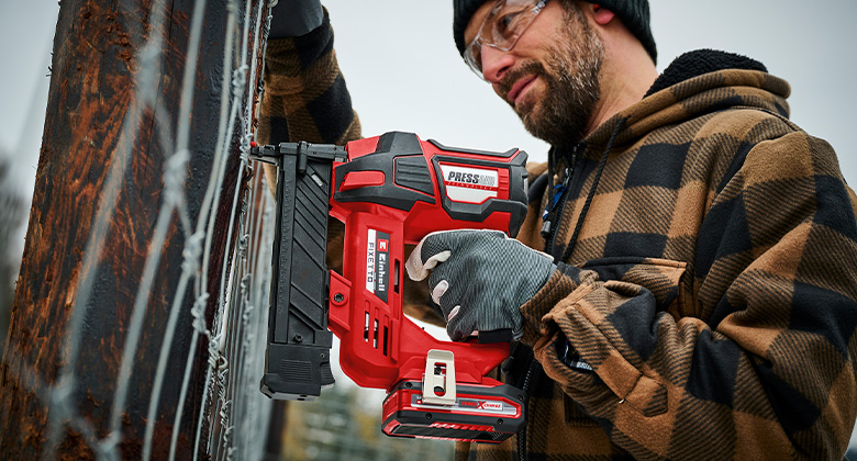 Ein Mann arbeitet mit einem Einhell Akku-Tacker FIXETTO, um einen Draht an einen Zaun zu befestigen, dabei trägt er Handschuhe und eine Schutzbrille.