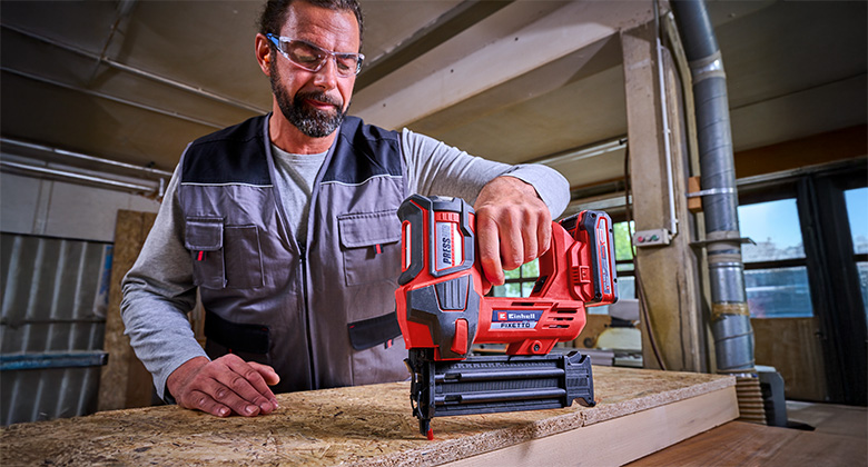 Ein Mann verwendet einen Einhell Akku-Nagler FIXETTO, um ein Holzstück zu bearbeiten, während er Schutzbrille und Arbeitskleidung trägt.