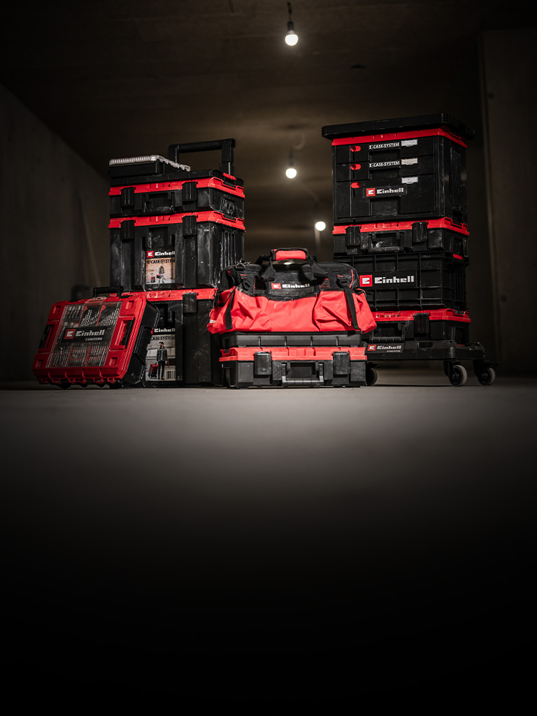 Several black and red Einhell E-Case toolboxes and bags are stacked and arranged in a dark room.
