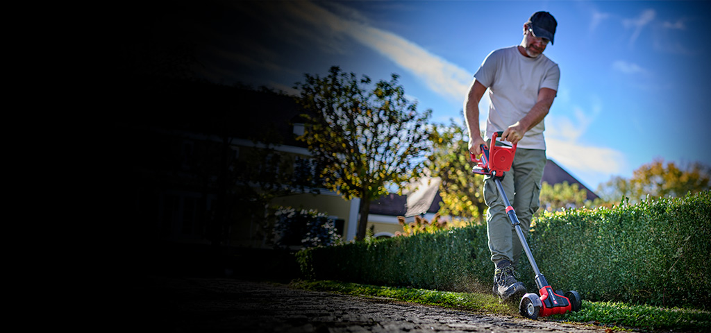 Ein Mann reinigt mit dem Einhell Akku-Fugenreiniger Pflasterfugen entlang einer Hecke im Garten.