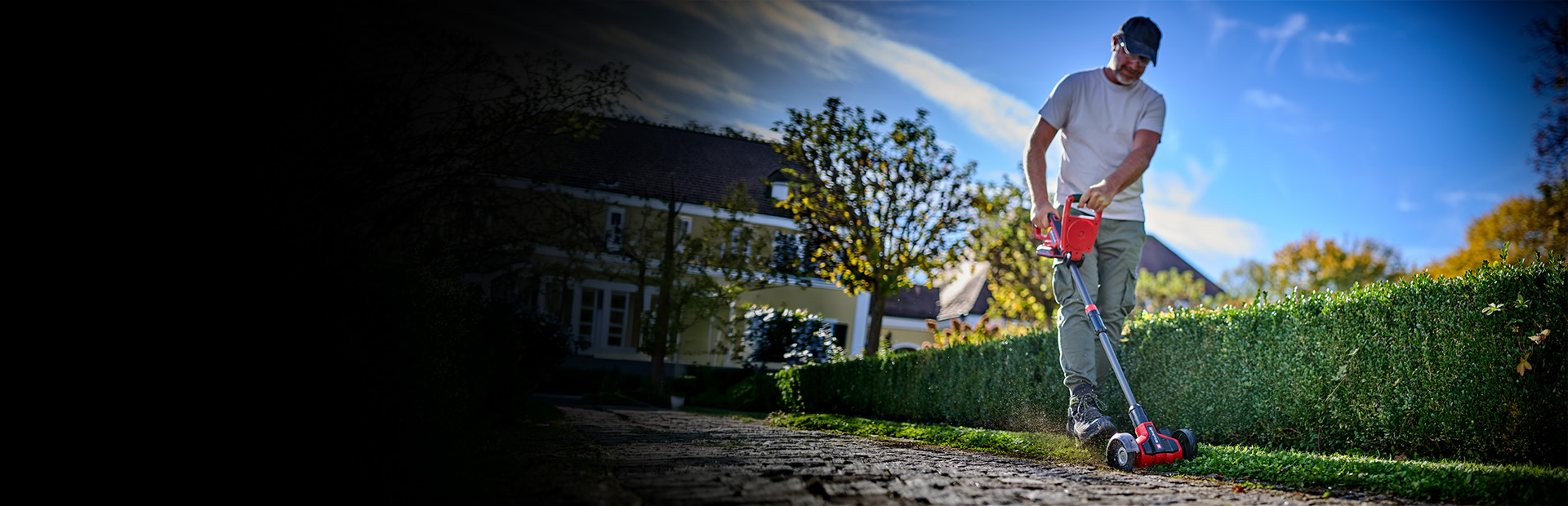 Ein Mann reinigt mit dem Einhell Akku-Fugenreiniger Pflasterfugen entlang einer Hecke im Garten.