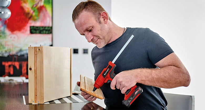 Man applies adhesive to a wooden board with a red cordless glue gun and holds another board in his hand.