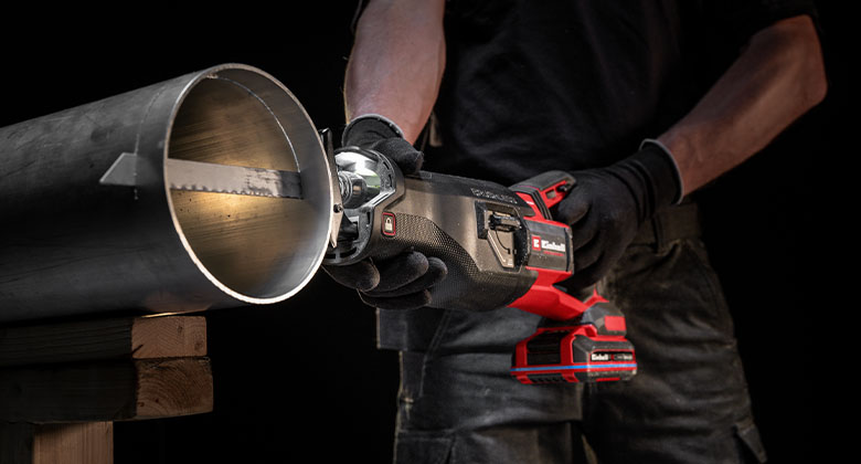 Person wearing gloves cutting a metal pipe with a red cordless reciprocating saw on a wooden support.