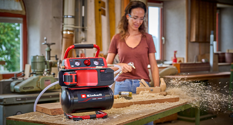 A woman works in a workshop with a red Einhell cordless compressor as wood chips scatter.