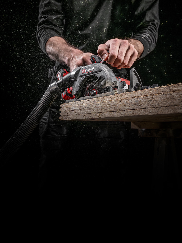 Person guides an Einhell circular saw with vacuum hose through a wooden board, sawdust flying