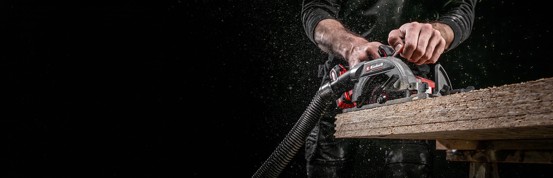 Person guides an Einhell circular saw with vacuum hose through a wooden board, sawdust flying