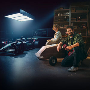 A father and child in a workshop with a homemade soapbox car. In the background, a Mercedes-AMG PETRONAS Formula One car and Einhell tools.