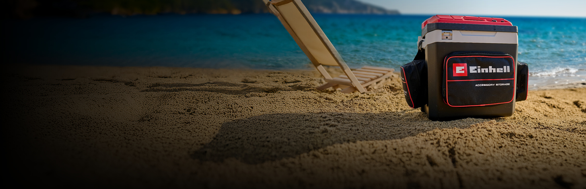The picture shows an Einhell cool box on the beach next to a lounger. In the background you can see the blue sea and a coastal landscape.