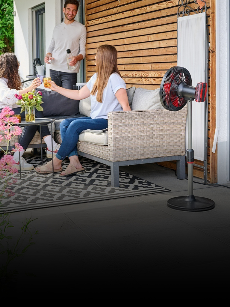 Three people enjoy drinks on a cozy patio surrounded by greenery and flowers. A red and black Einhell cordless fan is placed beside the seating area, providing airflow on a sunny day.