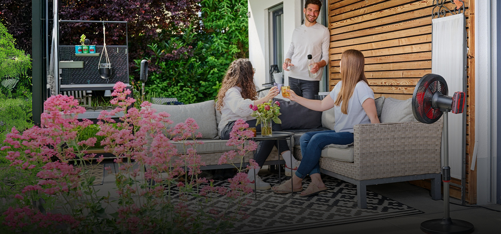 Three people enjoy drinks on a cozy patio surrounded by greenery and flowers. A red and black Einhell cordless fan is placed beside the seating area, providing airflow on a sunny day.