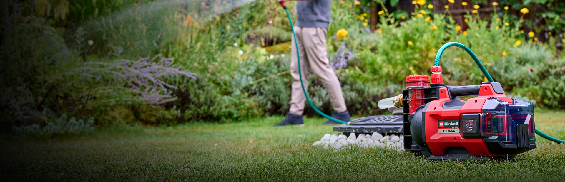 A man in casual clothing waters his garden with a hose, connected to a red and black Einhell cordless water pump placed on the grass in the foreground. The background shows lush green plants and yellow flowers.
