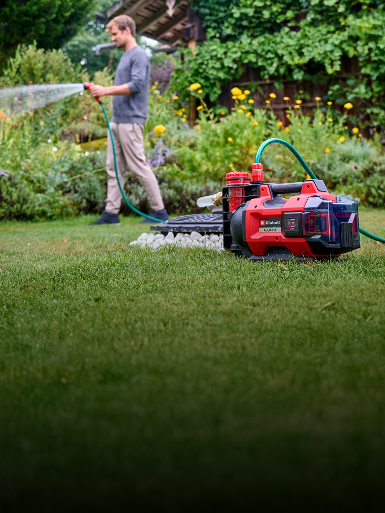 A man in casual clothing waters his garden with a hose, connected to a red and black Einhell cordless water pump placed on the grass in the foreground. The background shows lush green plants and yellow flowers.