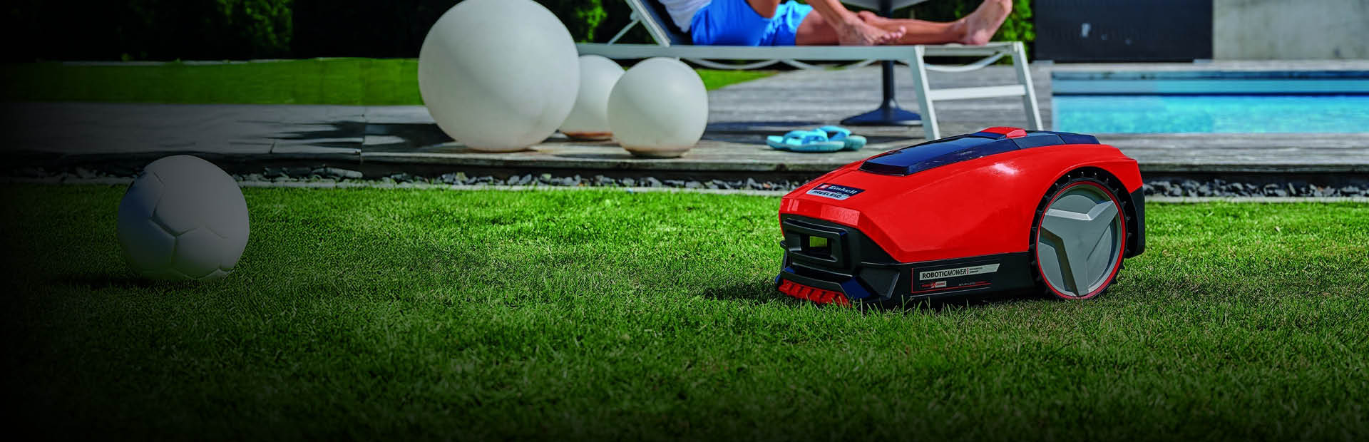 A man relaxes on a sun lounger near a swimming pool while a red robotic lawn mower autonomously trims the grass in the foreground.