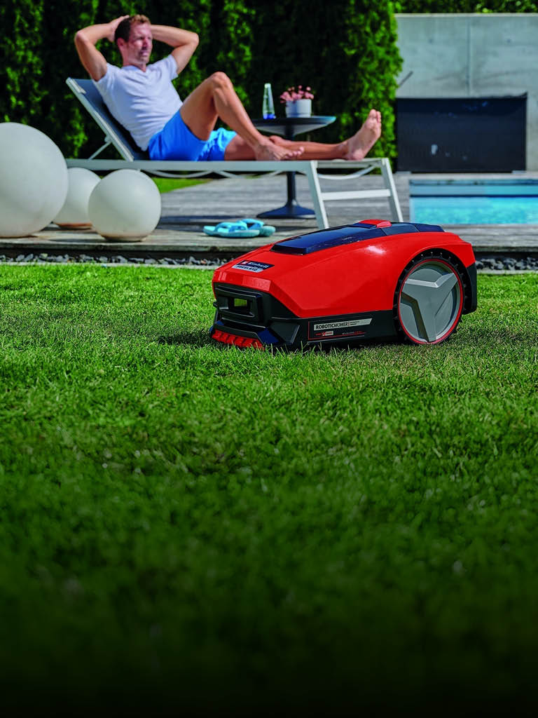 A man relaxes on a sun lounger near a swimming pool while a red robotic lawn mower autonomously trims the grass in the foreground.