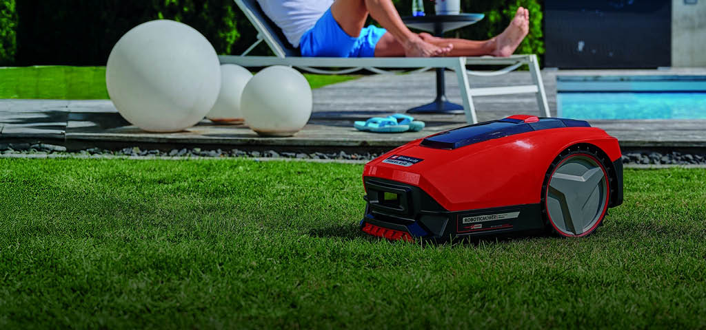 A man relaxes on a sun lounger near a swimming pool while a red robotic lawn mower autonomously trims the grass in the foreground.