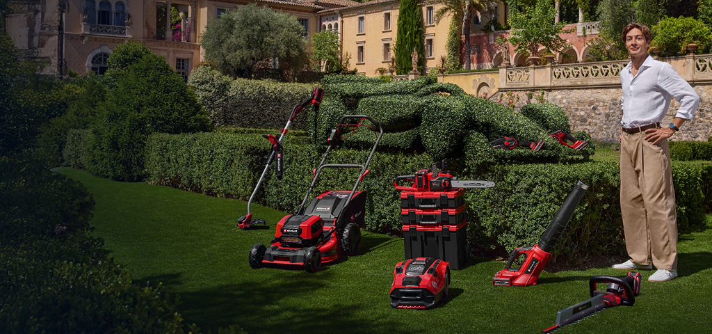 George Russell stands in a Mediterranean garden with Einhell cordless garden tools. Next to him, a race car-shaped hedge. 