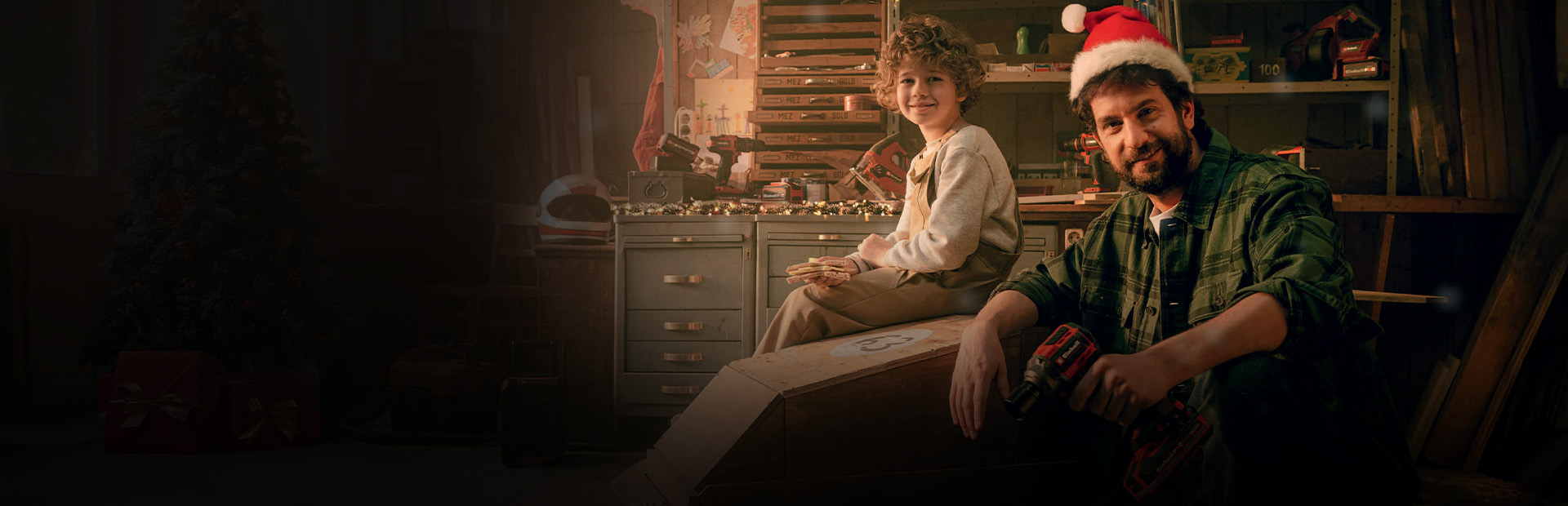 A child sits on a wooden car, while a man in a Santa hat kneels next to it holding a cordless screwdriver. In the background is a workbench, and a red-wrapped gift lies nearby.