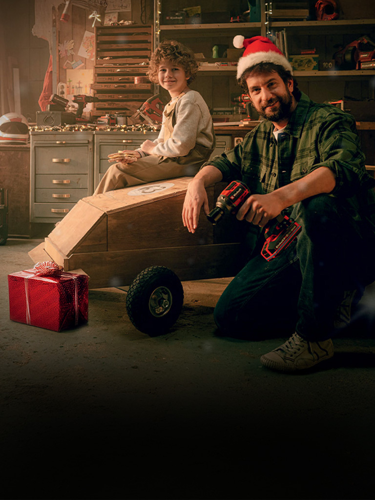 A child sits on a wooden car, while a man in a Santa hat kneels next to it holding a cordless screwdriver. In the background is a workbench, and a red-wrapped gift lies nearby.