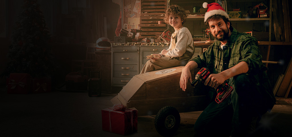 A child sits on a wooden car, while a man in a Santa hat kneels next to it holding a cordless screwdriver. In the background is a workbench, and a red-wrapped gift lies nearby.