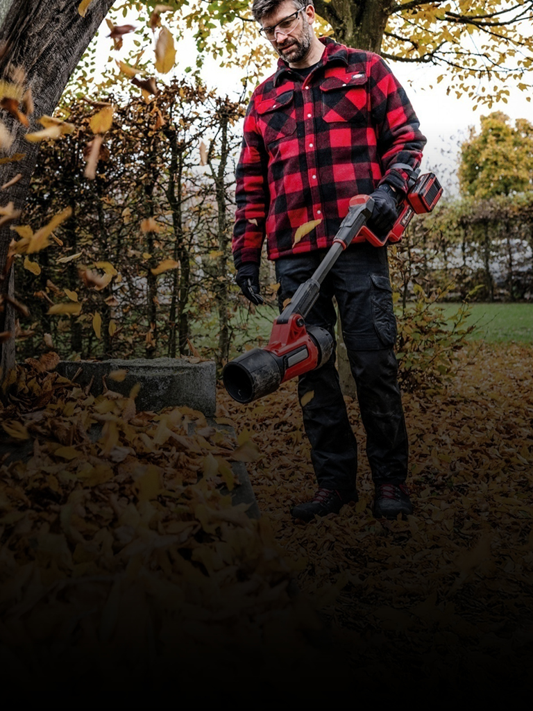 Das Bild zeigt eine Person, die im Herbst in einem Garten einen Einhell Laubbläser benutzt und eine rot-schwarz karierte Jacke, Handschuhe und eine Schutzbrille trägt.