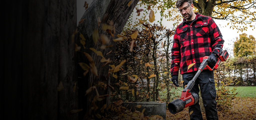 Das Bild zeigt eine Person, die im Herbst in einem Garten einen Einhell Laubbläser benutzt und eine rot-schwarz karierte Jacke, Handschuhe und eine Schutzbrille trägt.