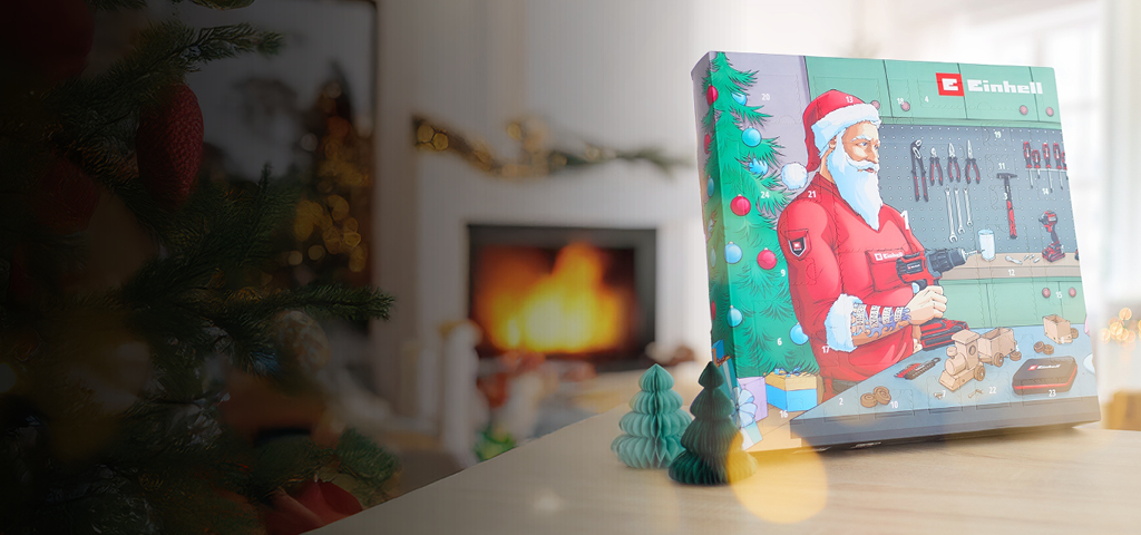 Das Bild zeigt eine gemütliche Weihnachtsszene mit einem geschmückten Baum, einem Kamin und einem Einhell-Adventskalender, auf dem der Weihnachtsmann in einer Werkstatt mit einer Bohrmaschine zu sehen ist.