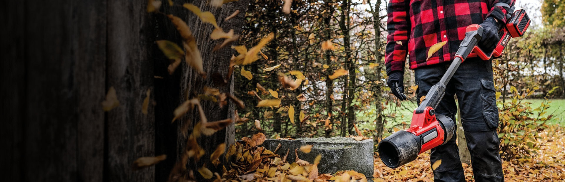 Eine Person in schwarzer Hose, Handschuhen und rot-schwarz karierter Jacke steht im Freien und hält einen roten, akkubetriebenen Laubbläser der Marke Einhell. Am Boden liegt herabgefallenes Herbstlaub, im Hintergrund sind Bäume zu sehen.