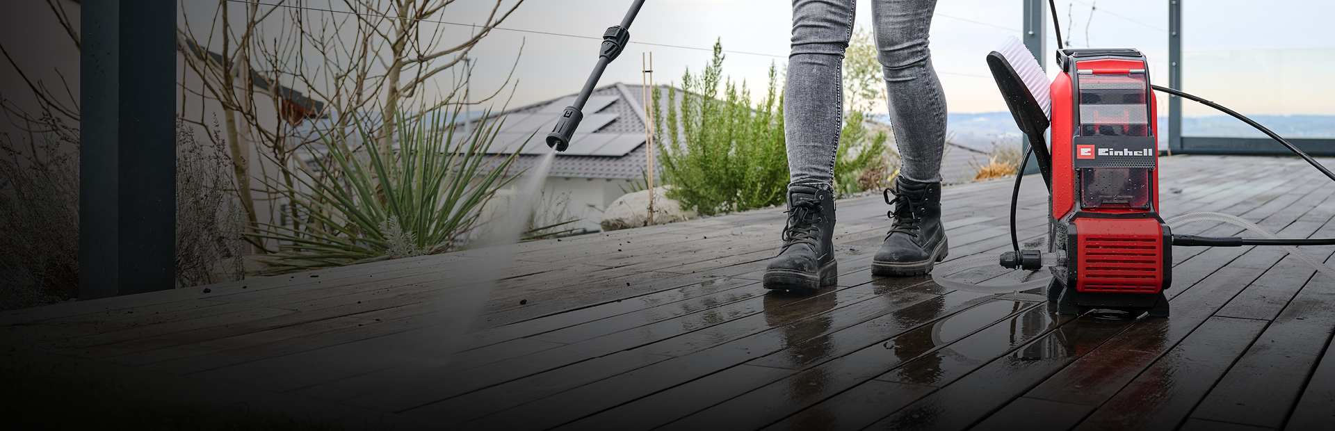 The image shows a person cleaning a wet wooden deck with a red Einhell pressure washer. Only their lower body is visible as they spray water across the surface outdoors.