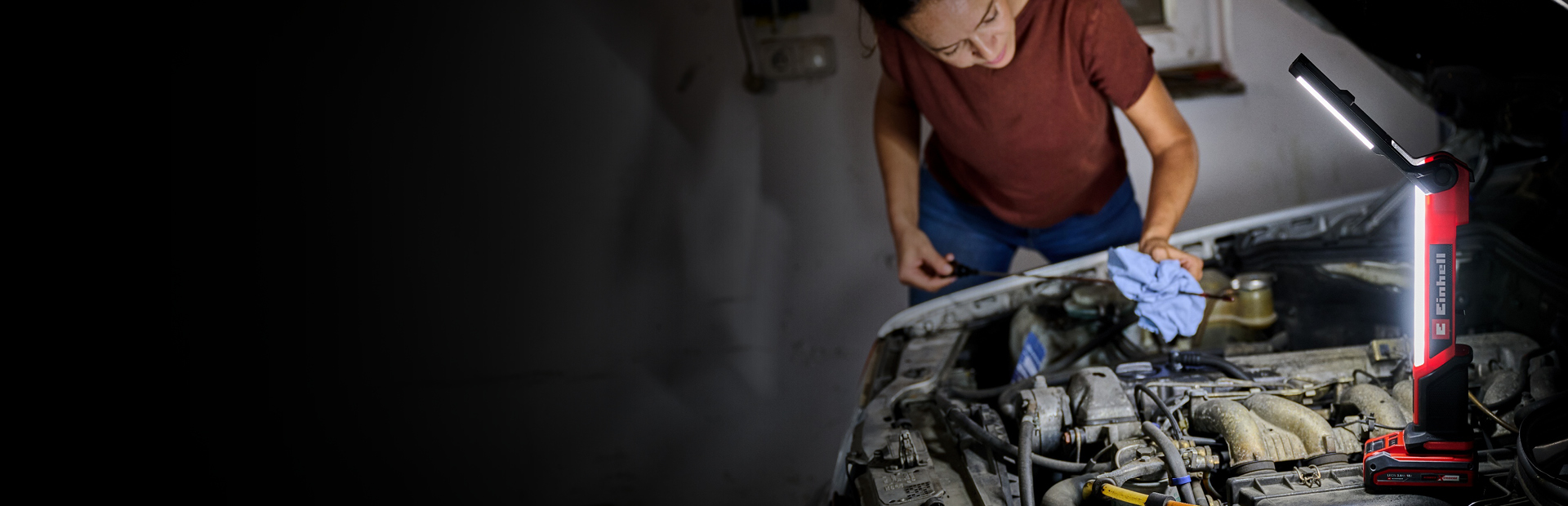 Das Bild zeigt eine Person, die in einer schwach beleuchteten Garage an einem Automotor arbeitet. Eine leuchtend rote und schwarze Einhell-Arbeitsleuchte beleuchtet den Motorbereich und sorgt für gute Sichtverhältnisse, während die Person ein Tuch und Werkzeuge verwendet.