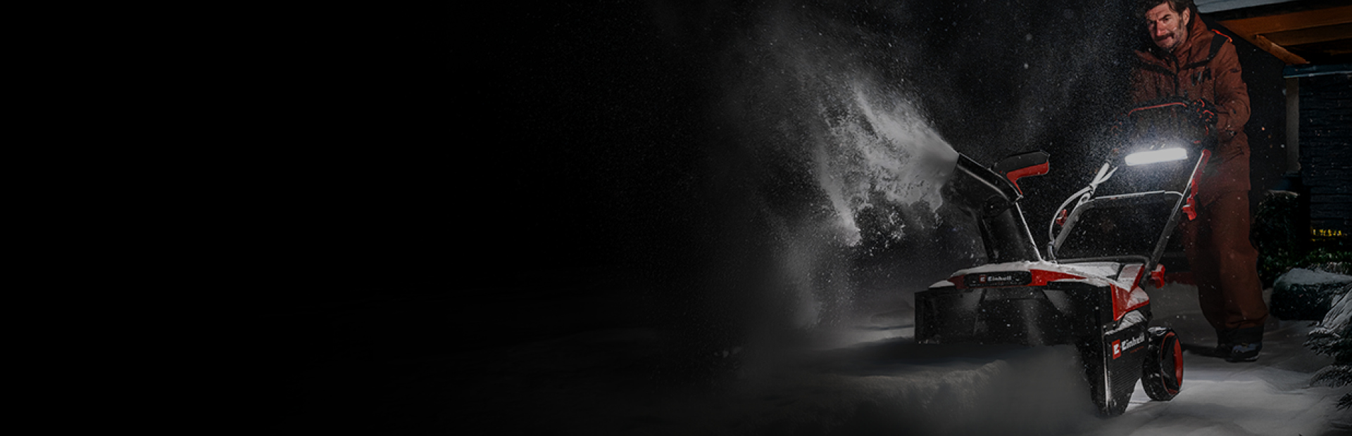 A man pushes a red-and-black snow thrower through deep snow at night.