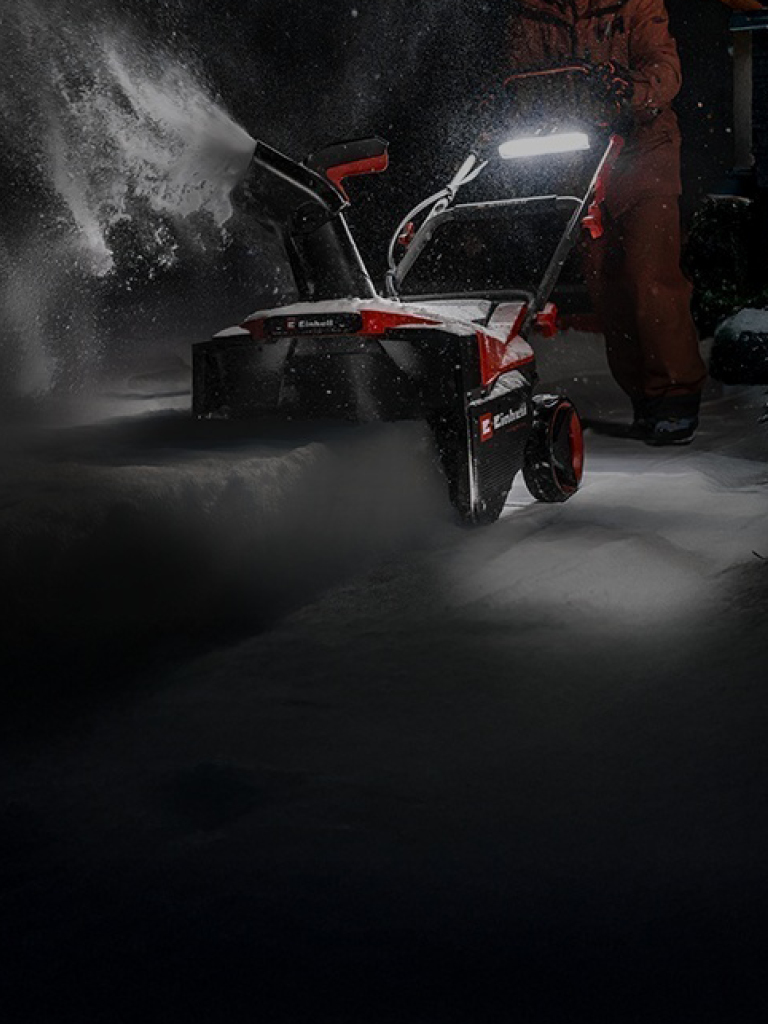 A man pushes a red-and-black snow thrower through deep snow at night.