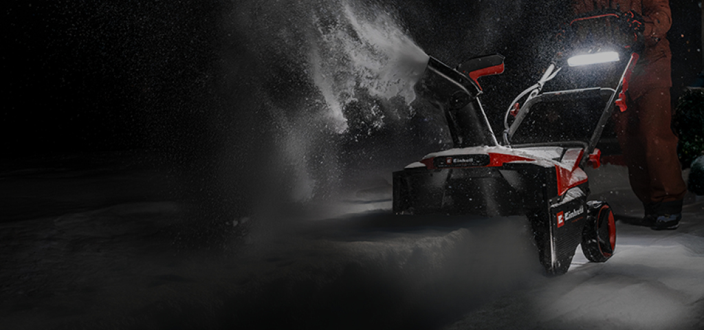 A man pushes a red-and-black snow thrower through deep snow at night.