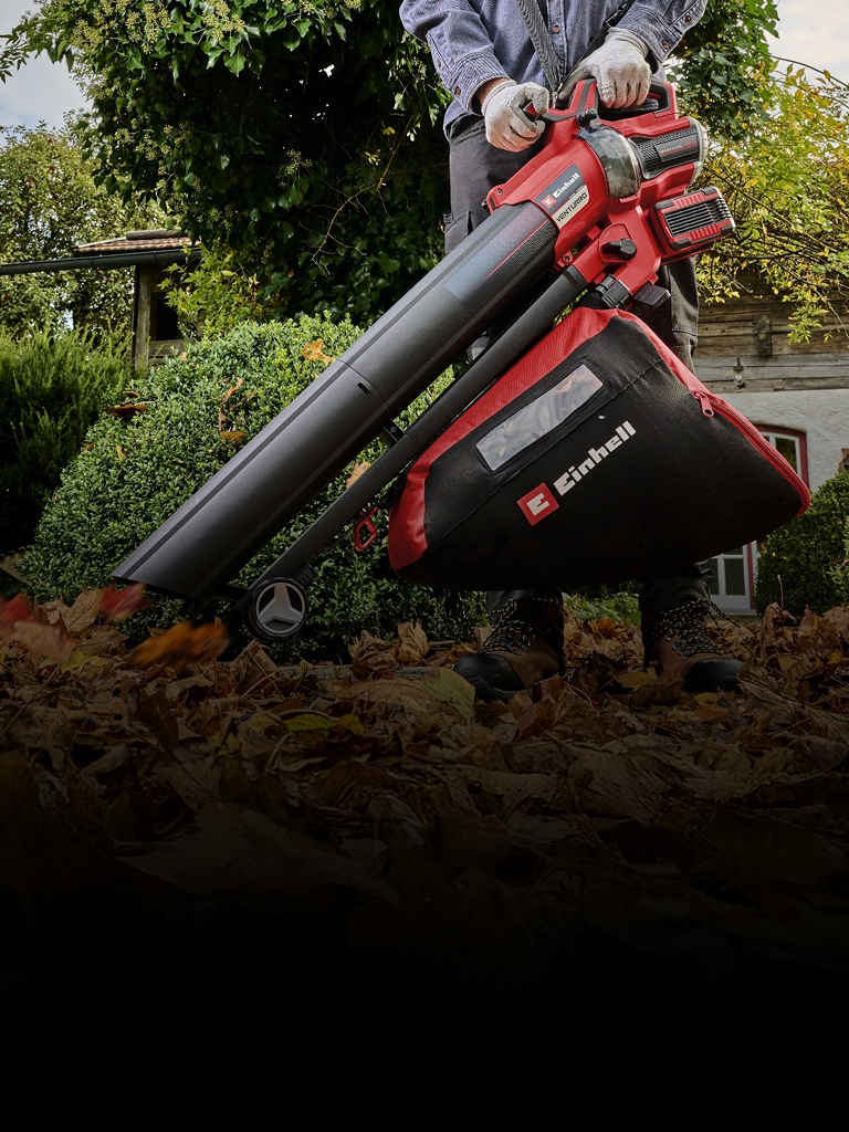 The image shows a person using a red and black Einhell leaf blower and vacuum to clear fallen leaves in a garden.