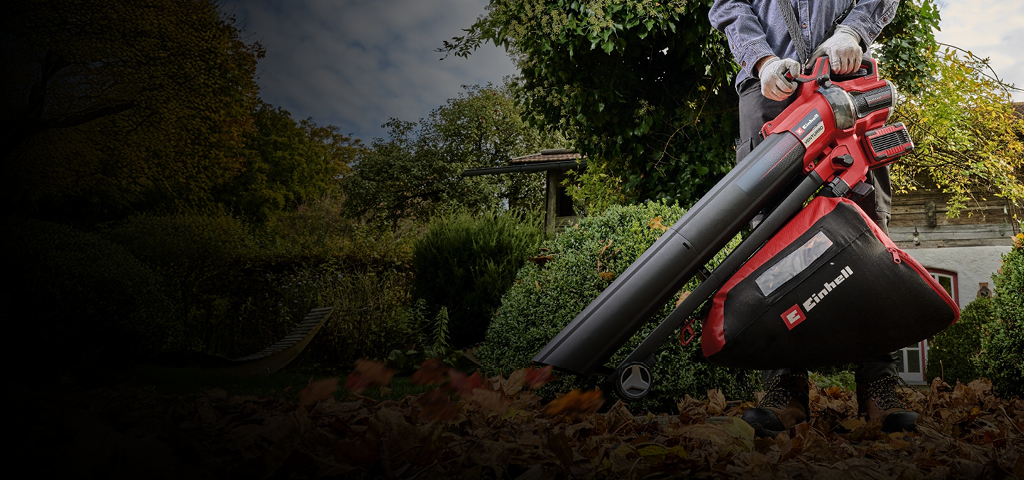 The image shows a person using a red and black Einhell leaf blower and vacuum to clear fallen leaves in a garden.