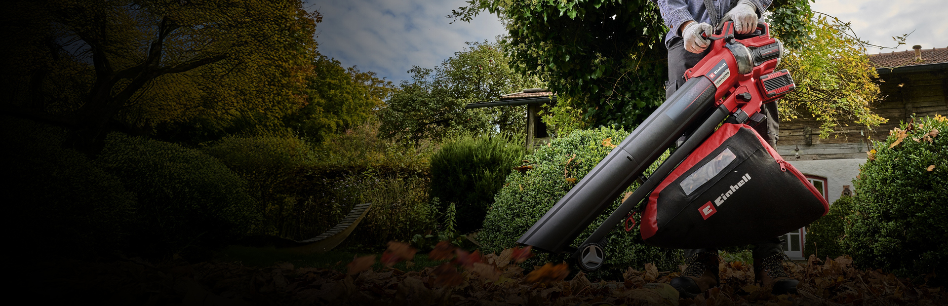 The image shows a person using a red and black Einhell leaf blower and vacuum to clear fallen leaves in a garden.