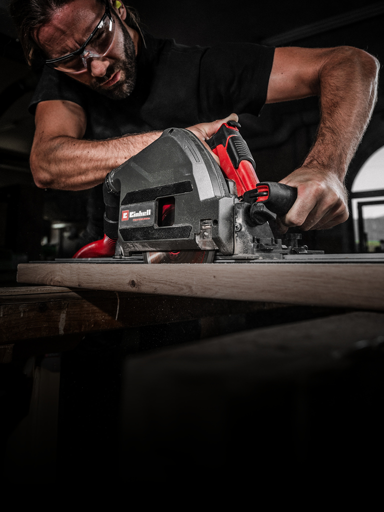A man wearing safety goggles operates a powerful Einhell circular saw to cut a wooden board in a workshop setting. His focused expression and firm grip emphasize precision and safety.