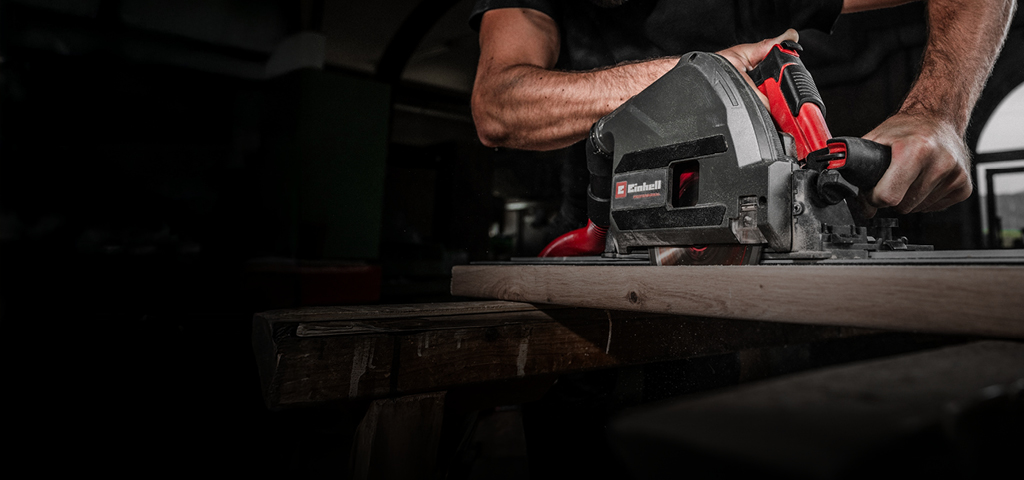 A man wearing safety goggles operates a powerful Einhell circular saw to cut a wooden board in a workshop setting. His focused expression and firm grip emphasize precision and safety.