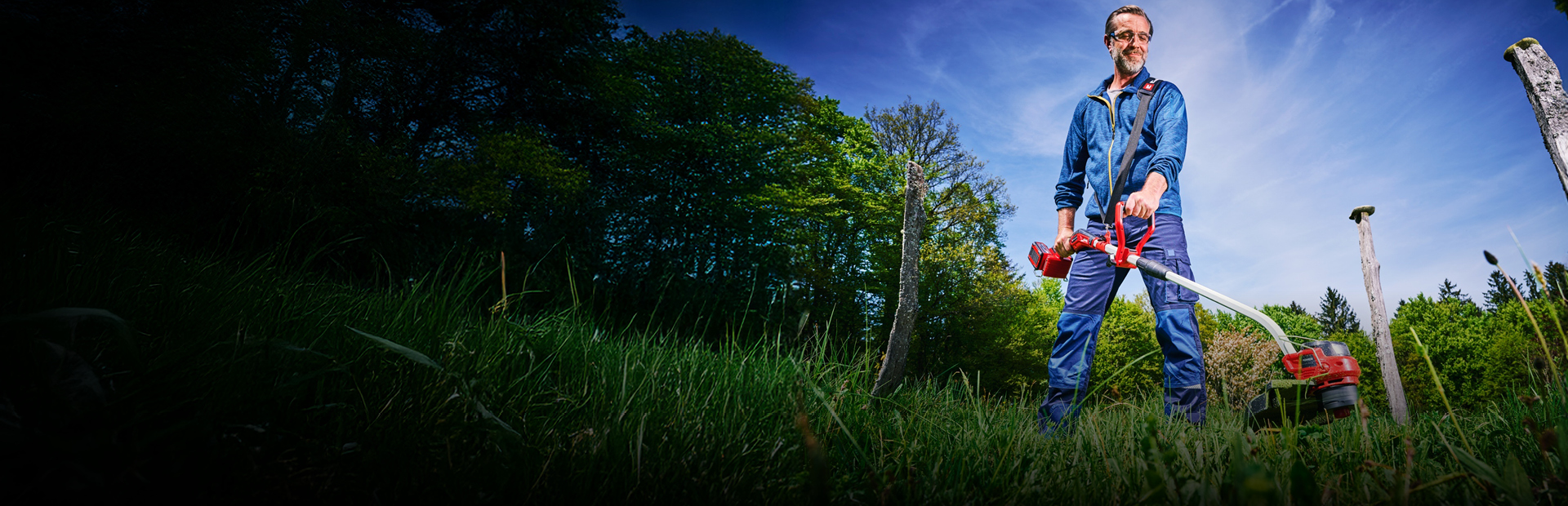 A man in blue work clothing and protection glasses, uses a red and black Einhell cordless lawn trimmer to trim tall grass between tree stumps in a green, wooded area.