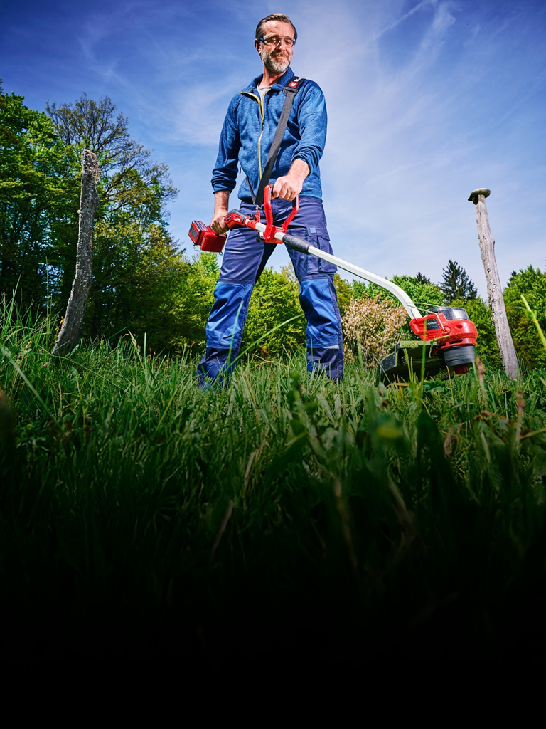 A man in blue work clothing and protection glasses, uses a red and black Einhell cordless lawn trimmer to trim tall grass between tree stumps in a green, wooded area.