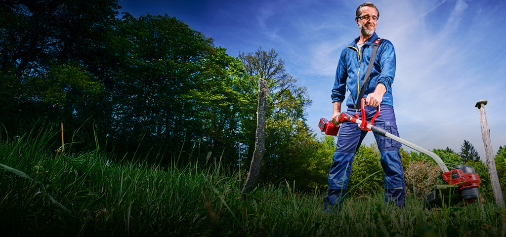 A man in blue work clothing and protection glasses, uses a red and black Einhell cordless lawn trimmer to trim tall grass between tree stumps in a green, wooded area.